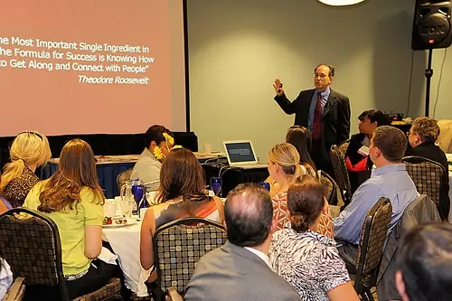 Arnold Sanow presenting to a seated audience beside a projected quote slide